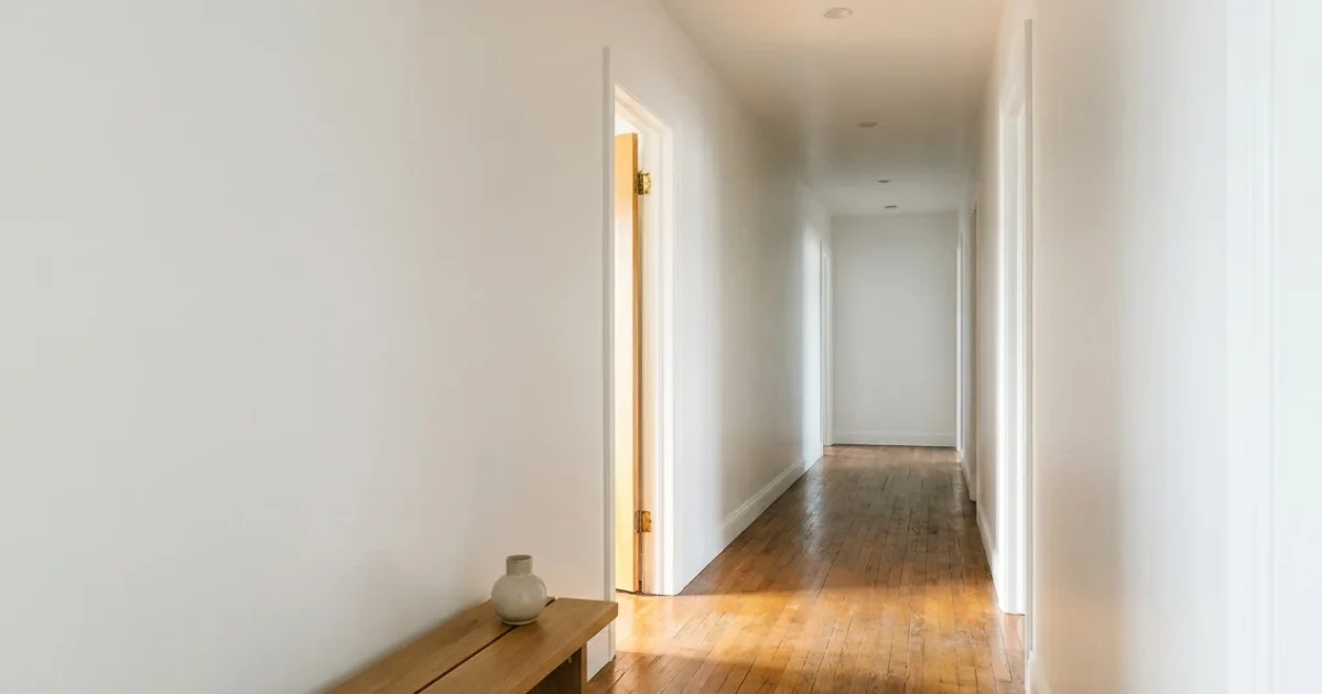 A bright, clean windowless hallway with white walls and hardwood floors, showing light reflecting from an adjacent room.
