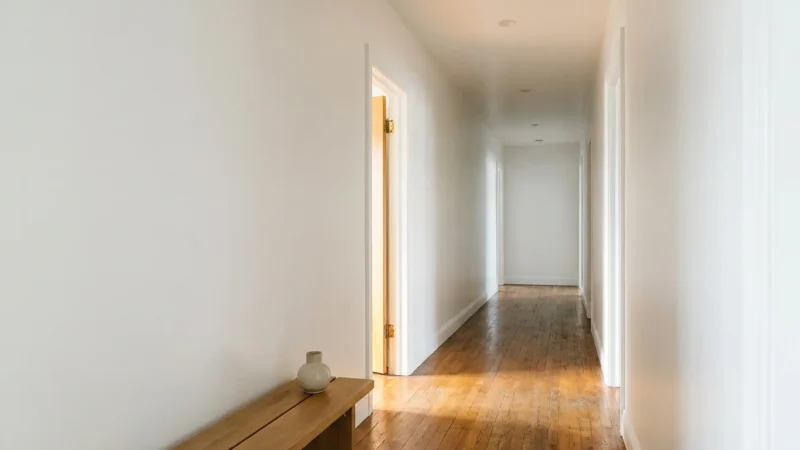 A bright, clean windowless hallway with white walls and hardwood floors, showing light reflecting from an adjacent room.