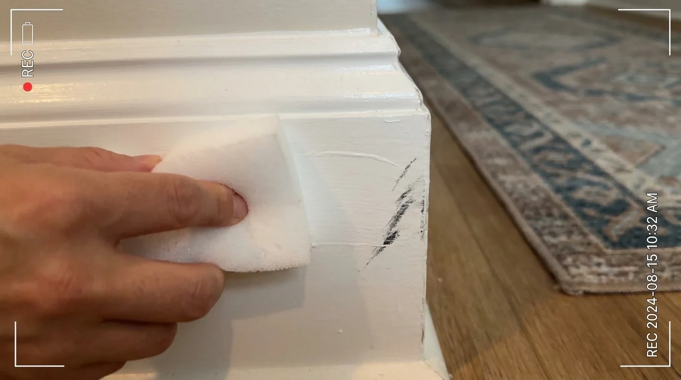 A close-up of a melamine sponge erasing a dark scuff mark from a white baseboard.
