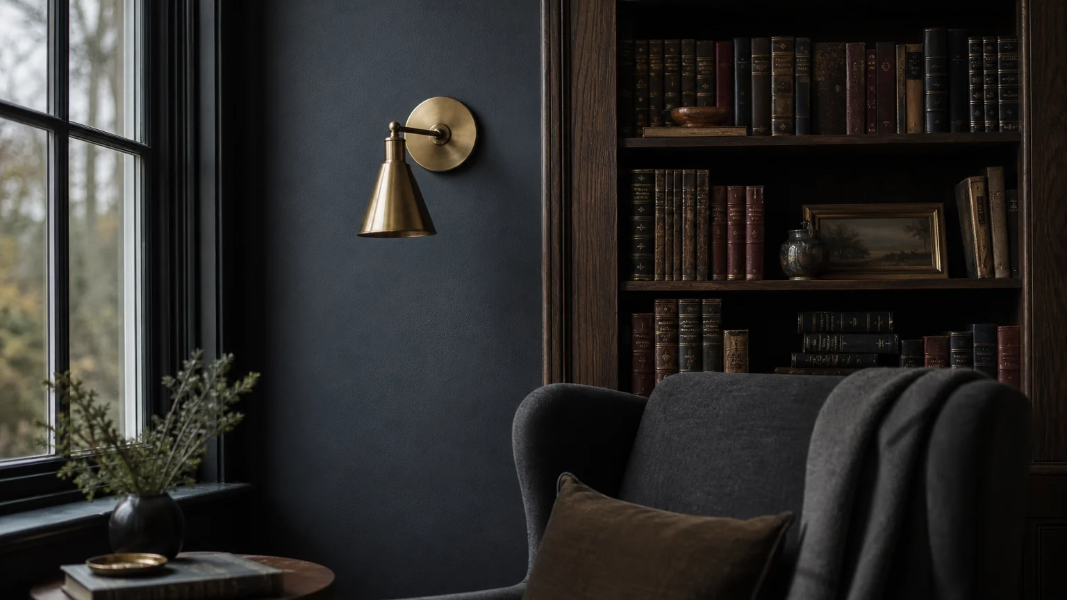 A moody slate blue library wall with a dark wood bookshelf and a brass sconce in soft daylight.