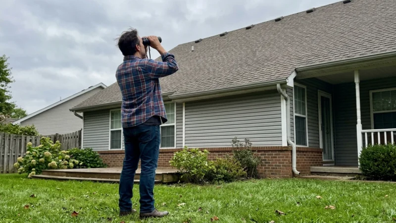 A homeowner in a flannel shirt looks up at a suburban house roof through binoculars on an overcast day.