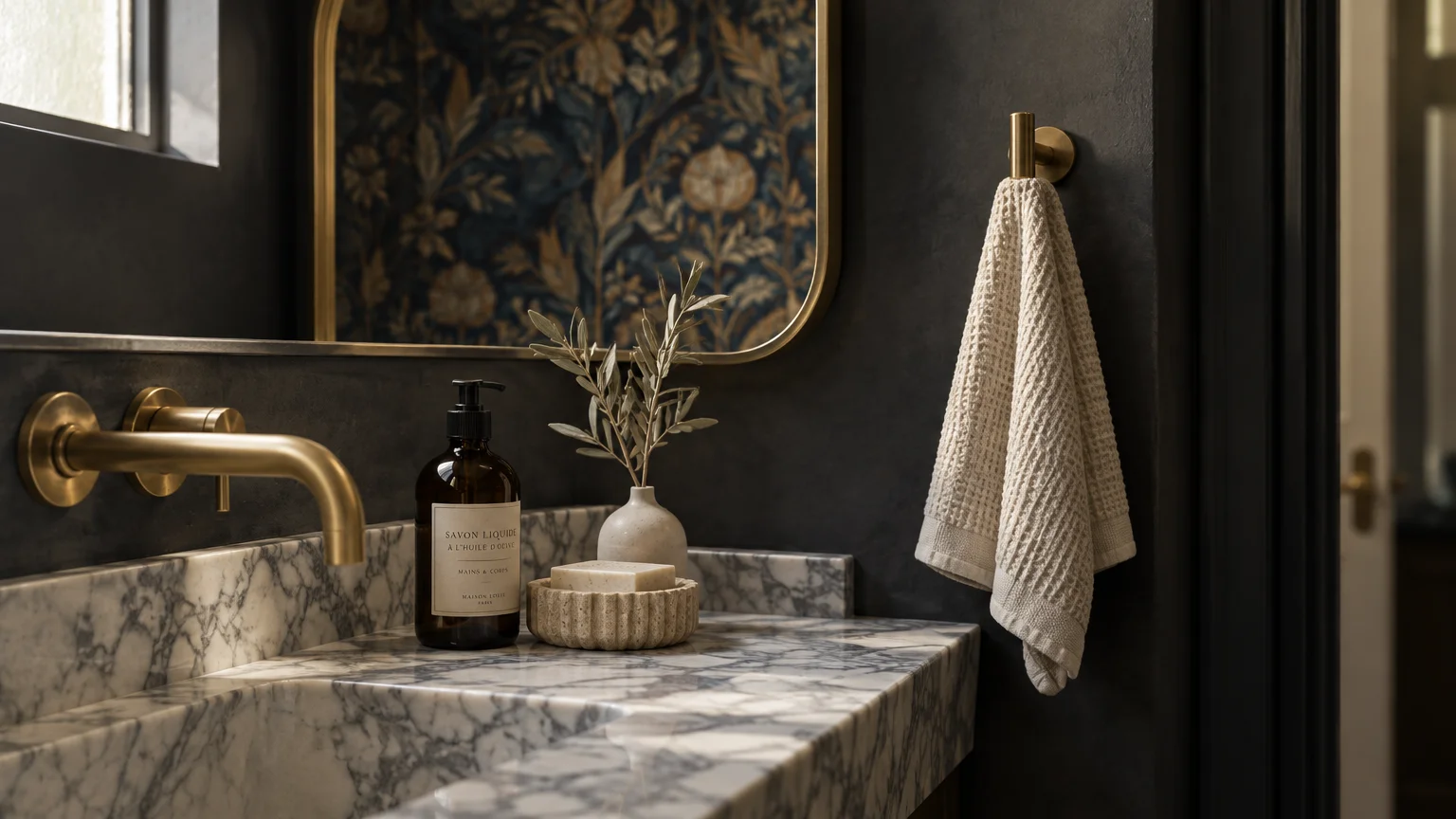 Marble vanity with brass wall-mount faucet, soap dispenser, small vase, brass-framed mirror, and waffle hand towel.