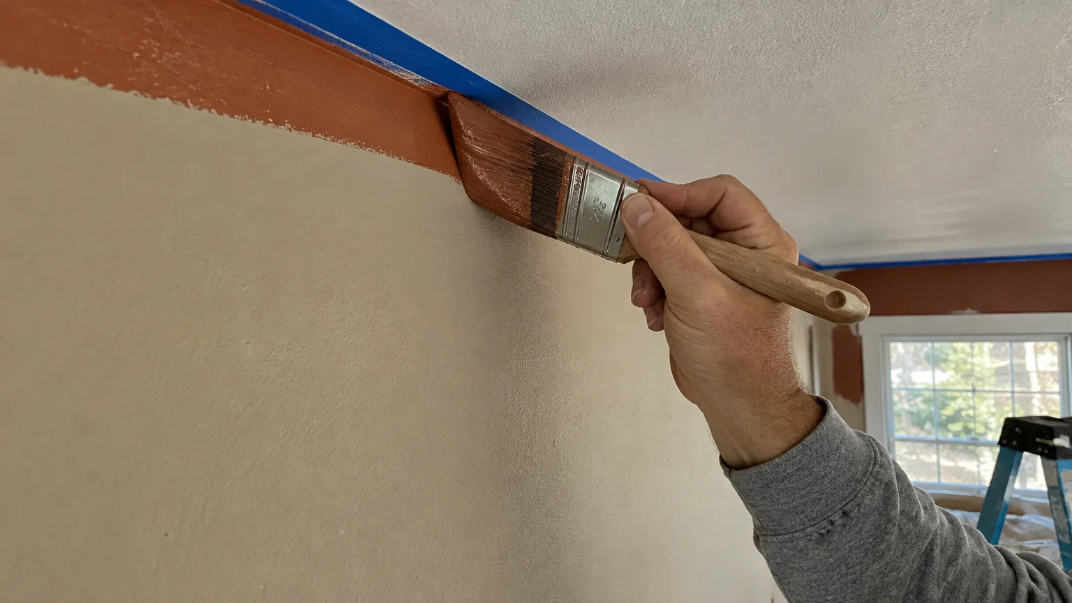 A person using an angled paintbrush to cut in warm brown paint along a taped ceiling line on a beige wall.