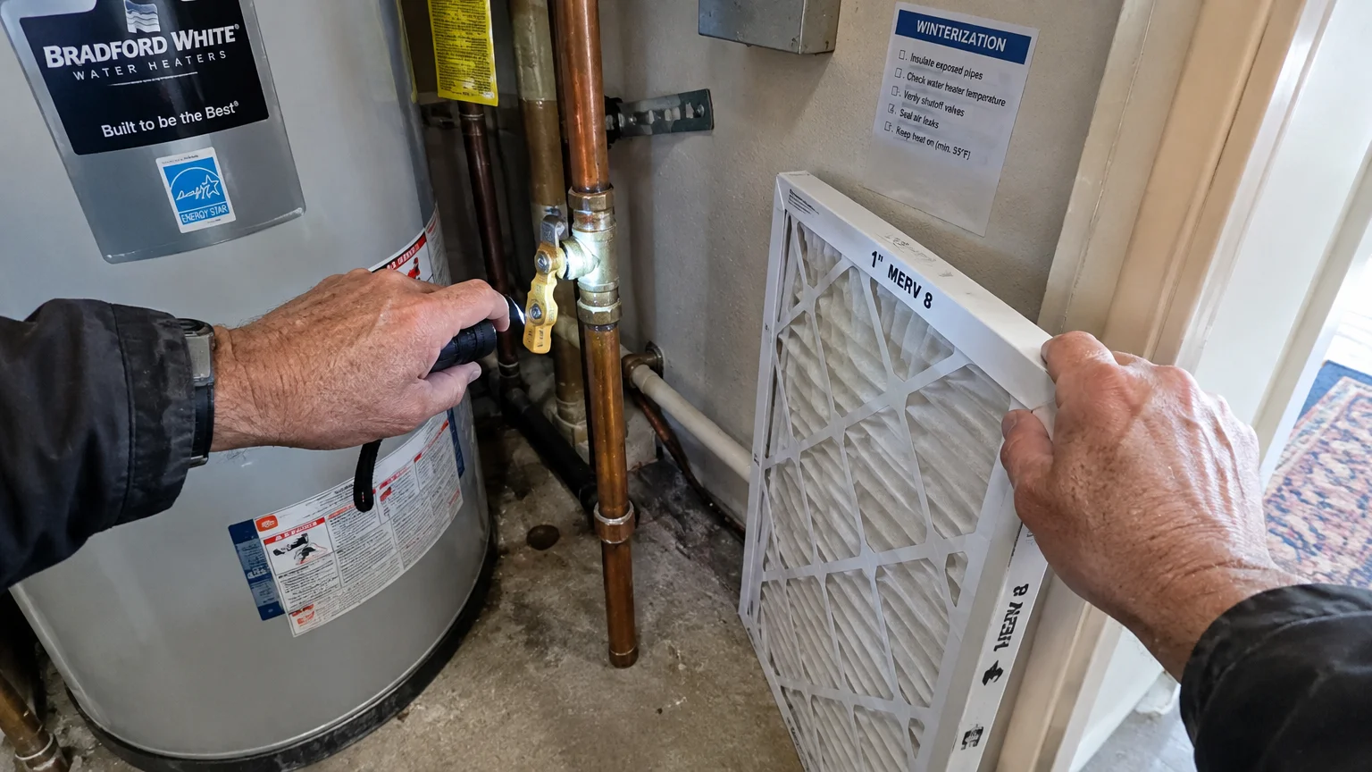 Hands holding a flashlight toward a water heater pipe valve next to a white furnace air filter.