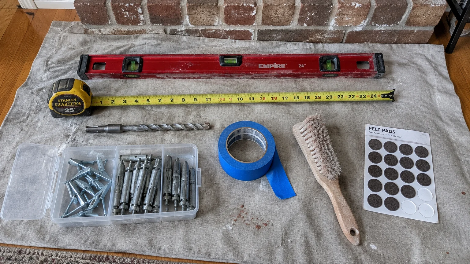 Level, extended tape measure, masonry drill bit, anchors in a plastic organizer, painter's tape, brush, and felt pads on cloth.