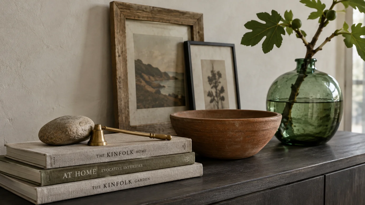 Framed art, stacked books with a brass bell, a terracotta bowl, and a green glass vase on a dark wood sideboard.